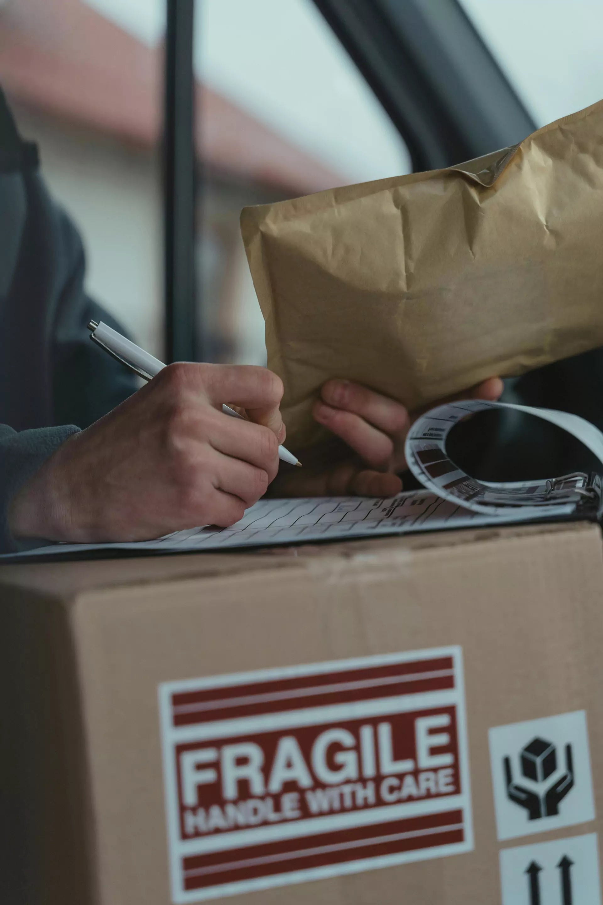 Person holding a brown envelope and completing paperwork on a box marked “Fragile.”